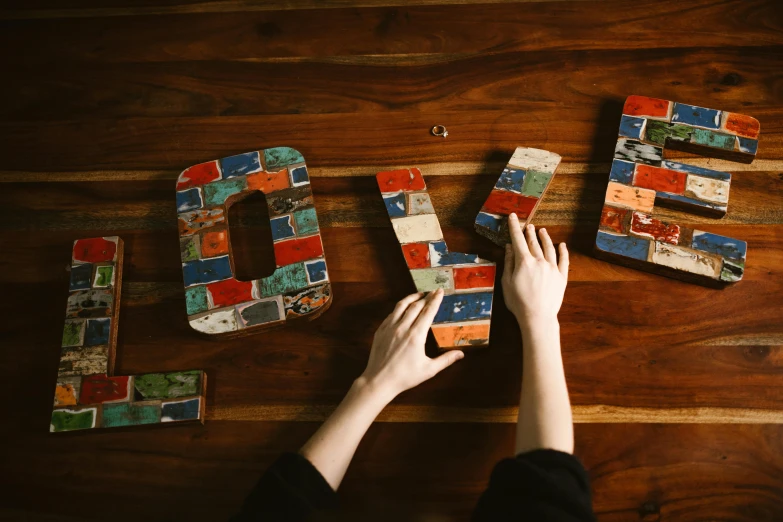 a person writing the word love on a wooden table, a mosaic, by Emma Andijewska, trending on unsplash, arts and crafts movement, brick, avatar image, enamel, shot on sony a 7