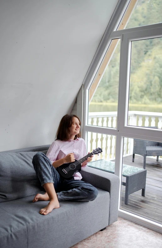 a woman sitting on a couch playing a guitar, lake house, cosy atmoshpere, teenager hangout spot, grey