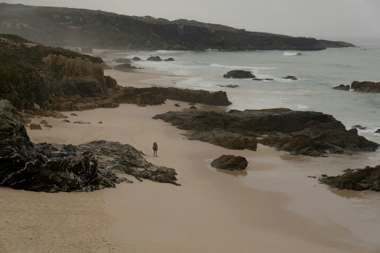 a man standing on top of a sandy beach next to the ocean, by Peter Churcher, pexels contest winner, portugal, rocky coast, walking down, slight overcast lighting