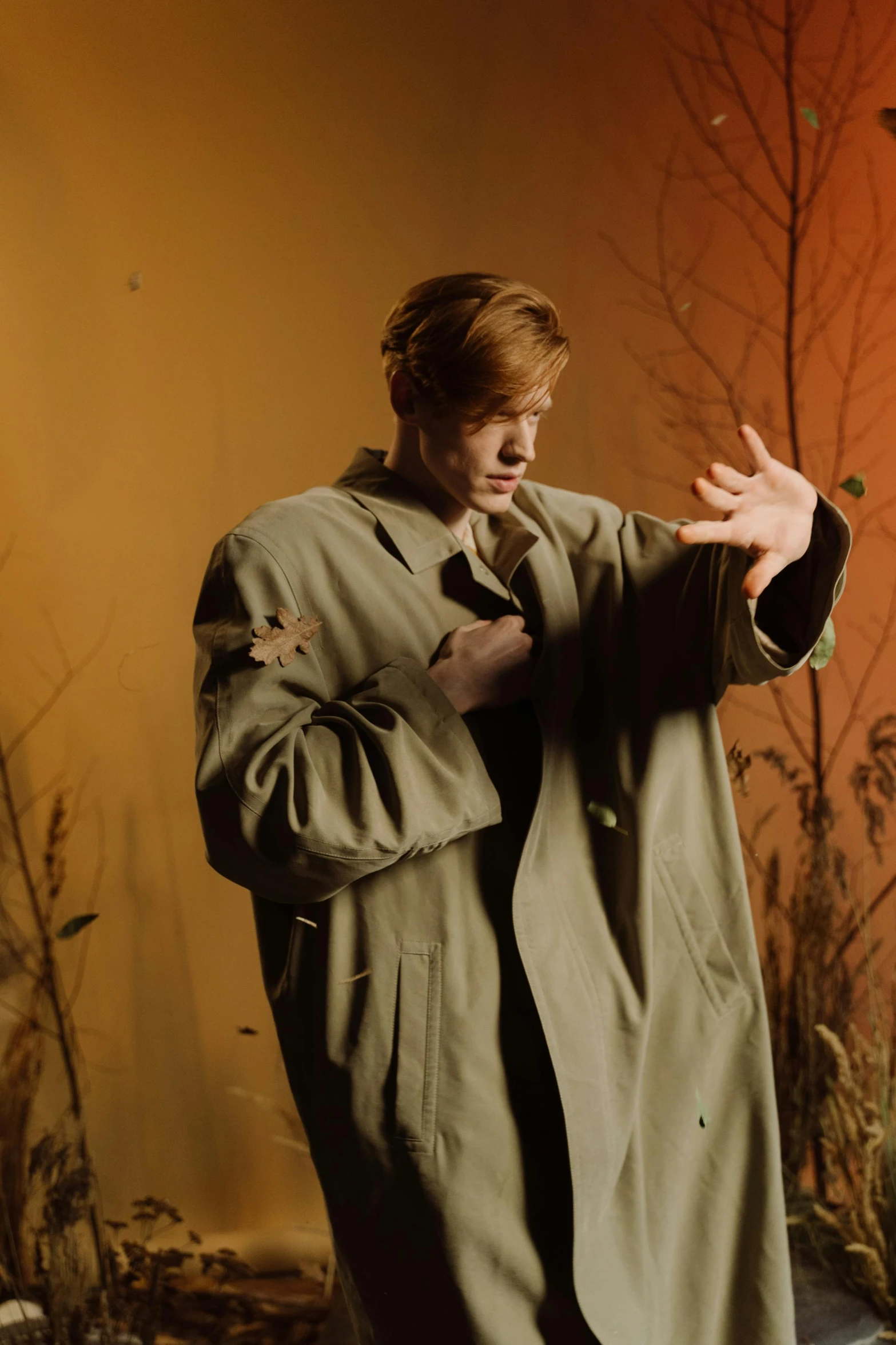 a man in a green coat standing next to a tree, an album cover, unsplash, realism, red haired teen boy, waving robe movement, standing in a dimly lit room, portrait photo of a backdrop