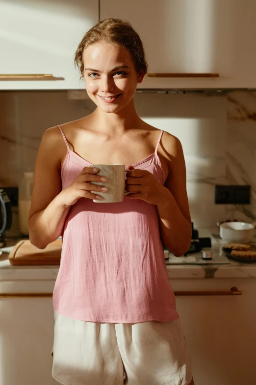 a woman standing in a kitchen holding a cup, pexels contest winner, renaissance, soft light 4 k in pink, sleeveless tops, wearing pajamas, warm smile