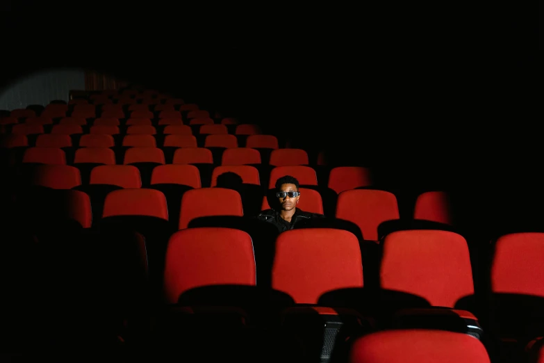 a man sitting in the middle of a row of red chairs, a picture, inspired by Elsa Bleda, pexels contest winner, portrait of willow smith, in a cinema, [ theatrical ], wong kar wai