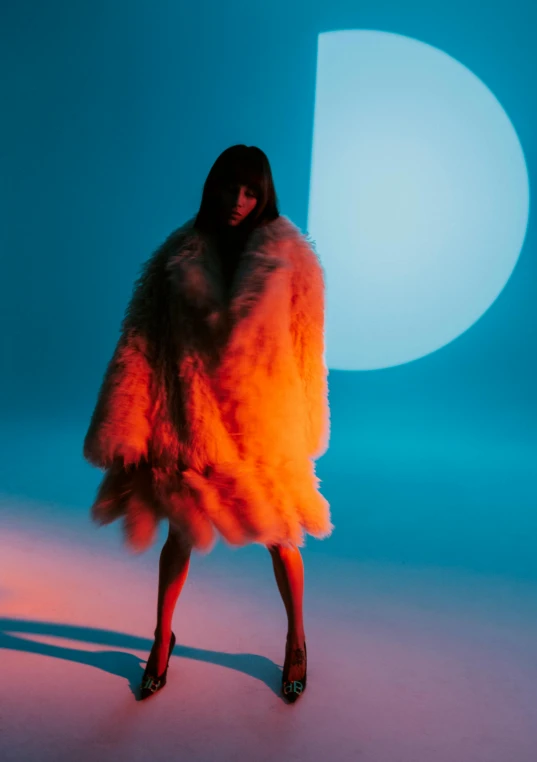 a woman in a fur coat standing in front of a blue background, an album cover, inspired by Elsa Bleda, orange reflective puffy coat, full body backlight, off - white collection, official music video