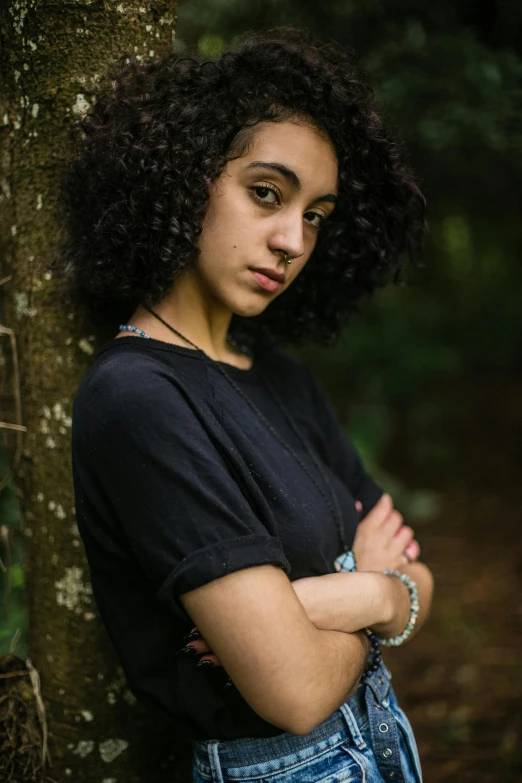 a woman leaning against a tree with her arms crossed, an album cover, by Lily Delissa Joseph, antipodeans, (dark shorter curly hair), ((portrait)), 18 years old, black in