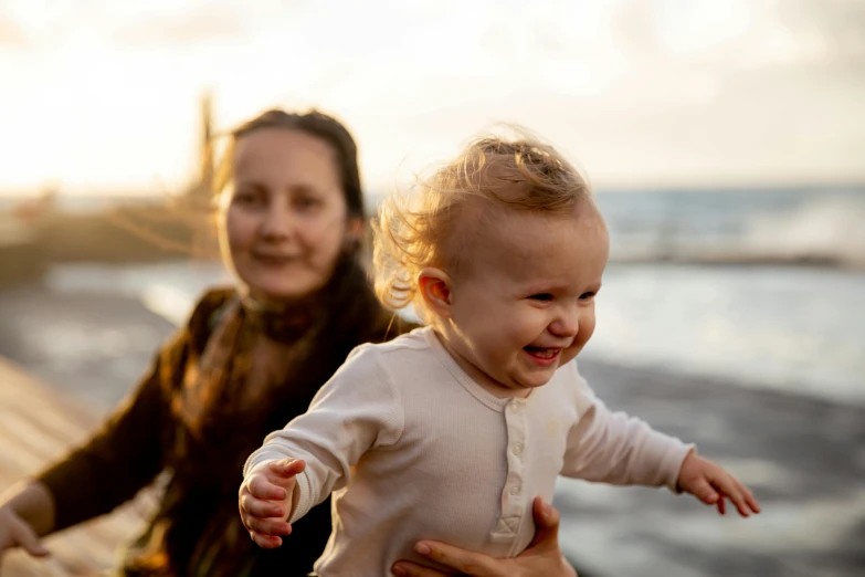 a woman holding a baby near a body of water, being delighted and cheerful, running towards camera, warm light, profile image