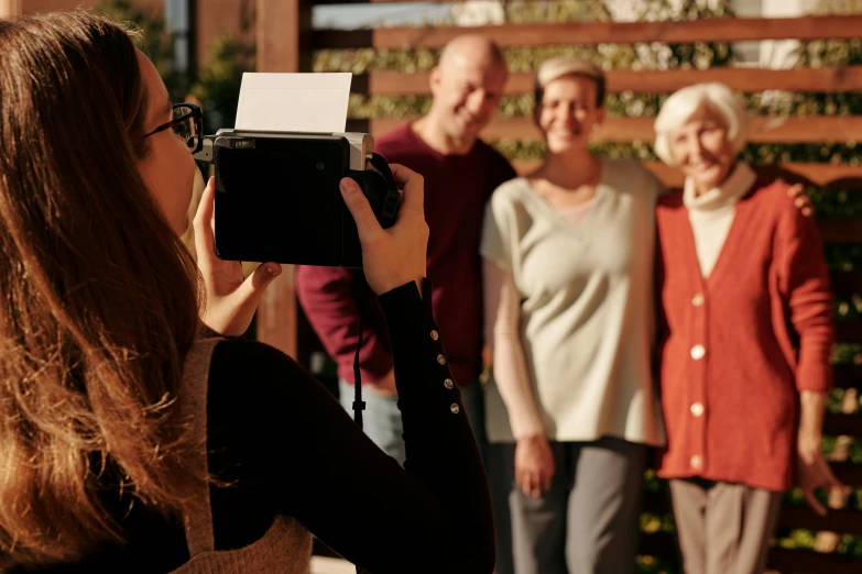 a woman taking a picture of a group of older people, moment cinebloom filter, in the sun, commercial photo shoot, holding polaroid camera