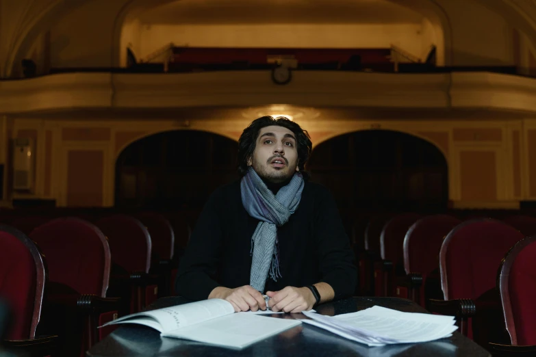 a man sitting at a table with papers in front of him, by Youssef Howayek, singing at a opera house, looking straight to camera, riyahd cassiem, threyda