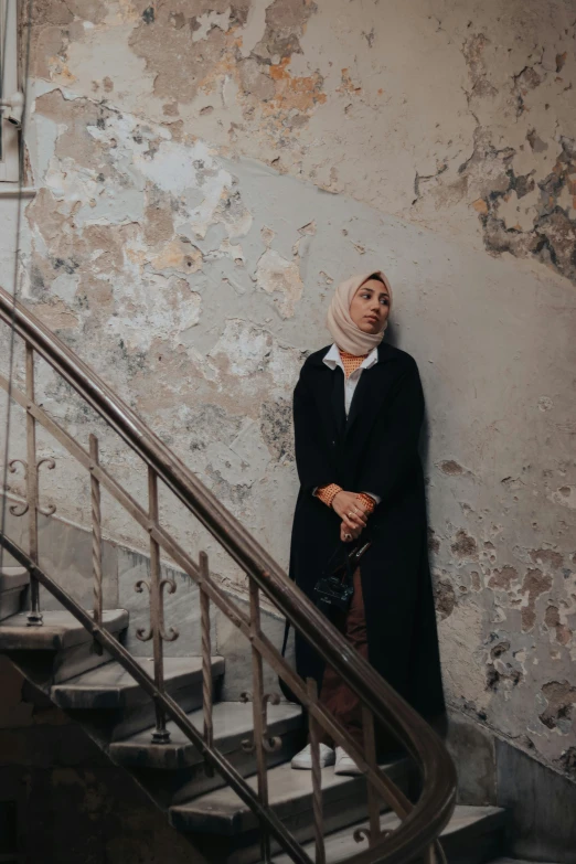 a woman standing next to a set of stairs, inspired by Modest Urgell, pexels contest winner, hurufiyya, hijab, distressed, ( ( theatrical ) ), leaning against the wall