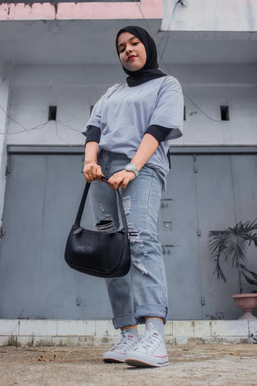 a woman standing in front of a building holding a purse, pexels contest winner, with ripped crop t - shirt, grey and dark theme, hijab, ( ( ( wearing jeans ) ) )