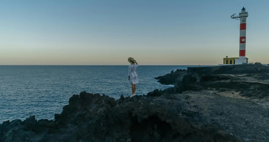 a woman standing on top of a cliff next to the ocean, by Alexis Grimou, happening, wearing a white bathing cap, dusk setting, high-quality photo, white lava