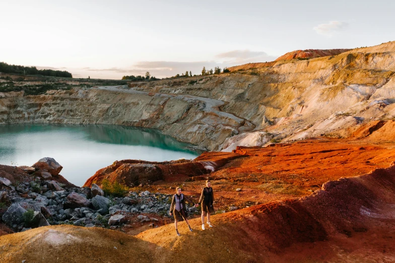 a couple of people standing on top of a hill next to a body of water, by Lee Loughridge, unsplash contest winner, in dusty open pit mine, vivid ember colors, manuka, blue and green and red tones