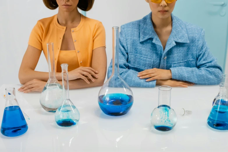 a couple of women sitting next to each other at a table, trending on pexels, analytical art, scientific glassware, blue clothing, acids, school class