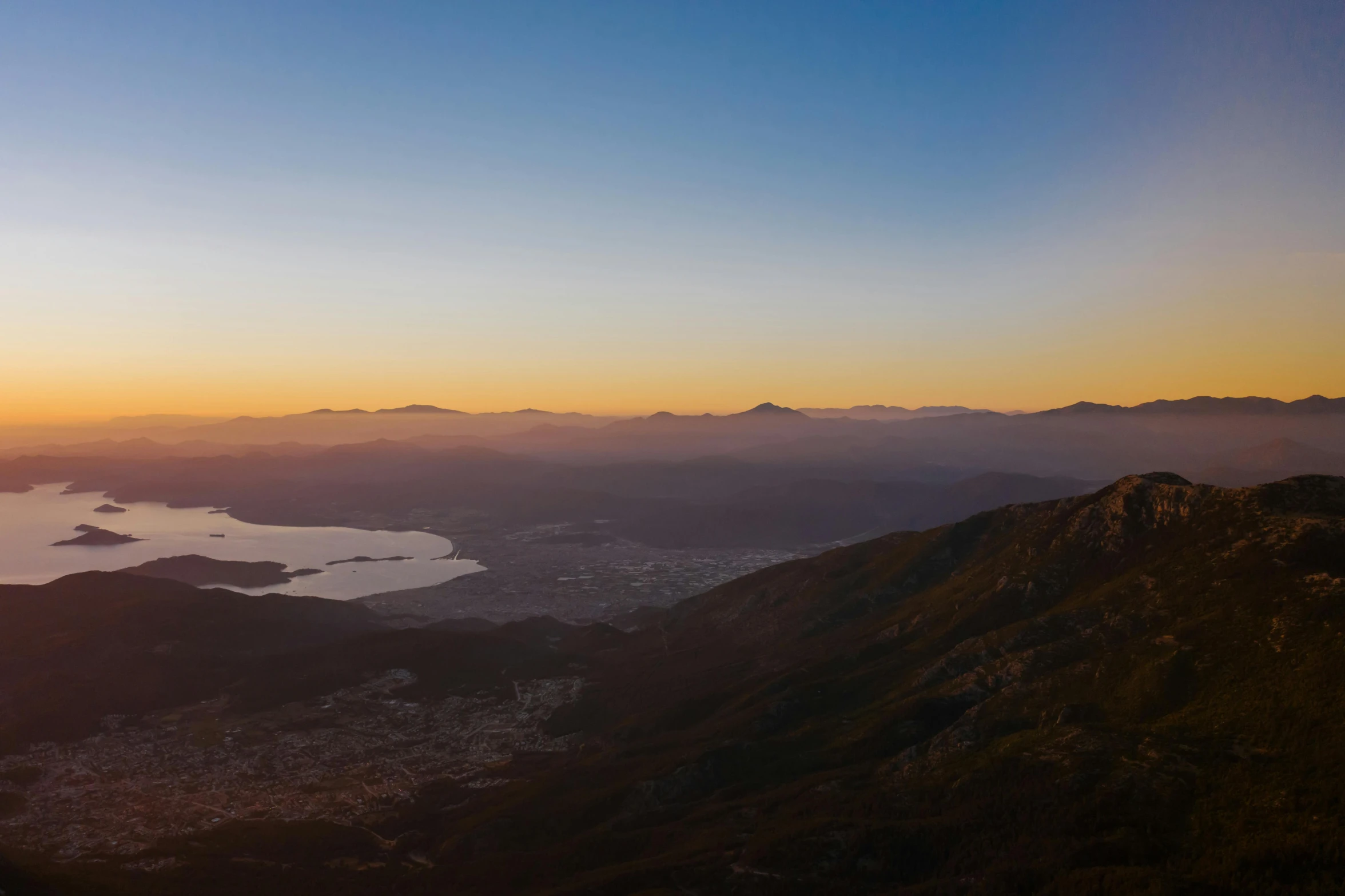 a view from the top of a mountain at sunset, mountains and lakes, sun and shadow over a city, golden hour photograph