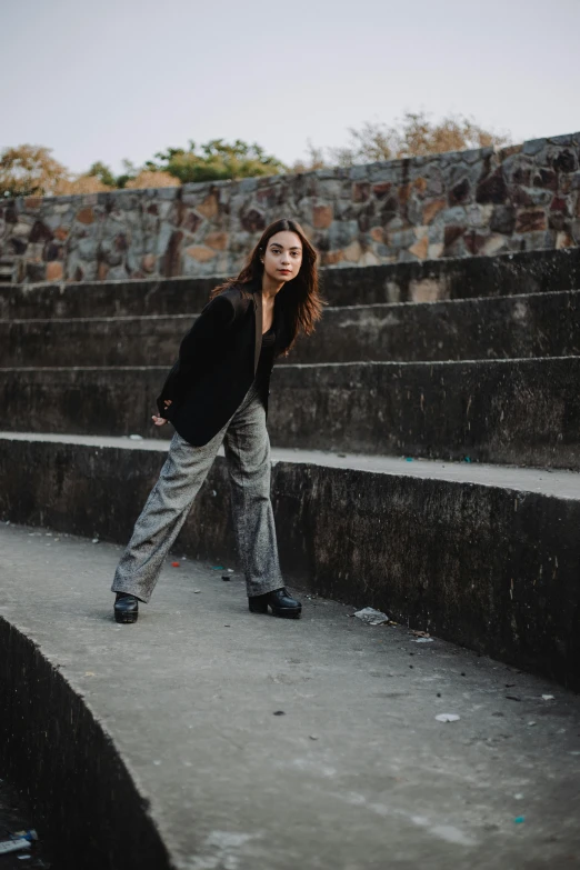 a woman standing in front of a stone wall, an album cover, pexels contest winner, grey pants and black dress shoes, in an arena pit, gongbi, wearing jacket