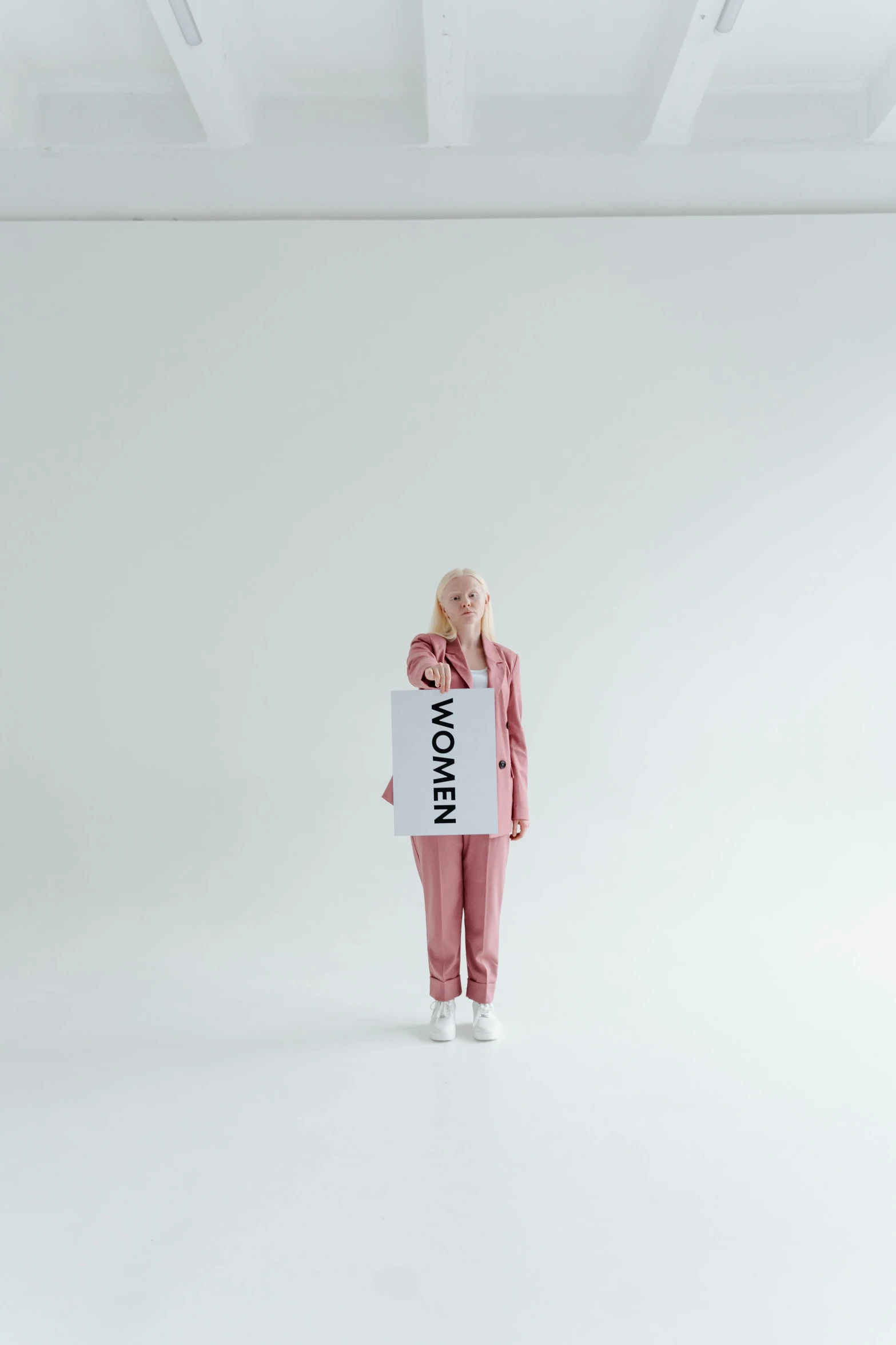 a woman standing in a white room holding a sign, an album cover, inspired by Louisa Matthíasdóttir, trending on unsplash, die antwoord yolandi visser, wearing track and field suit, healthcare, albino