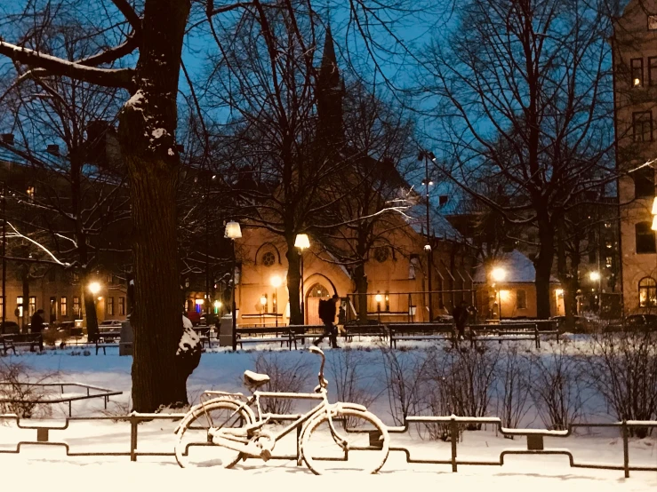 a bicycle parked in the middle of a snow covered park, by Jesper Knudsen, pexels contest winner, beautifully lit buildings, church in the background, people at night, swedish style