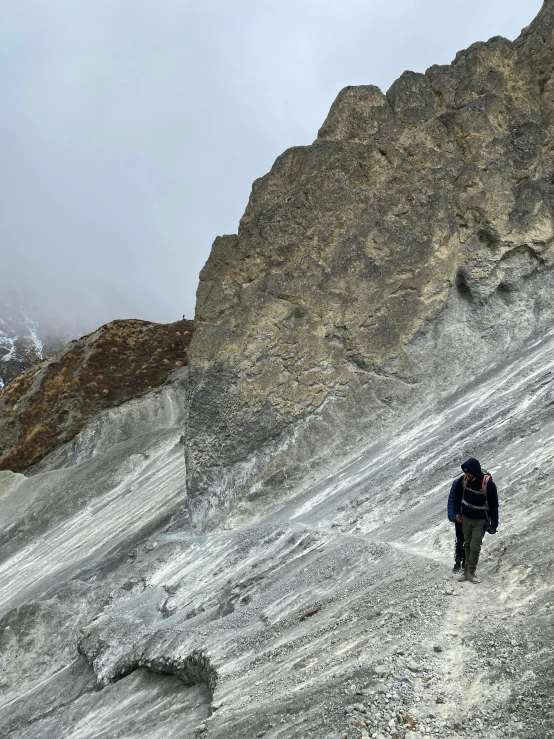 a man walking up the side of a snow covered mountain, with lots of dark grey rocks, covered in white flour, young himalayan woman, rock and dust