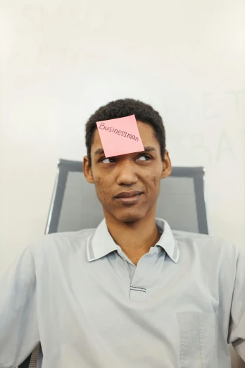 a man sitting in a chair with a post it on his forehead, an album cover, by Lily Delissa Joseph, unsplash, telegram sticker, serious business, pink face, taken in the late 2010s
