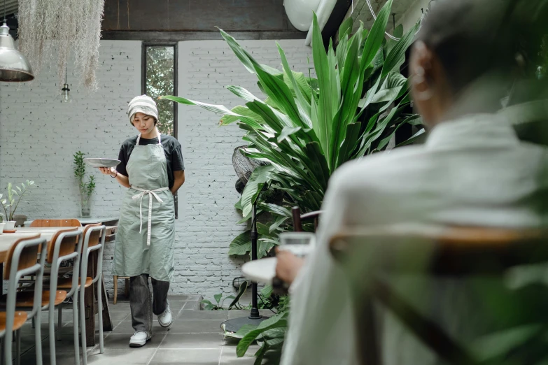 a woman that is standing in front of a table, trending on pexels, starbucks aprons and visors, large potted plant, cinematic medium shot, thumbnail