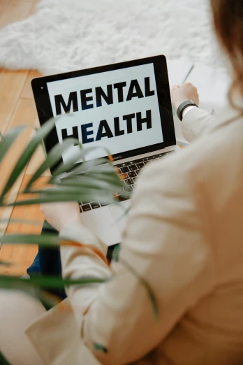 a woman sitting on the floor using a laptop with mental health written on the screen, zoomed in, thumbnail, shoulders and head, next to a plant