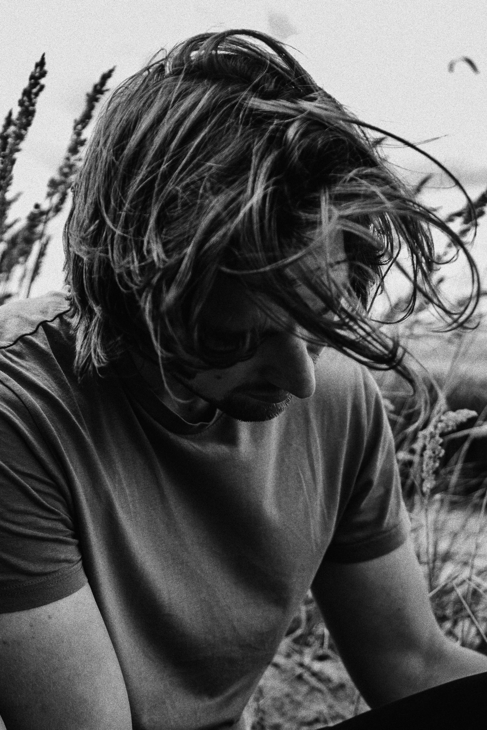 a black and white photo of a man sitting in a field, a black and white photo, unsplash, messy hair bedhead, on the coast, medium poly, bum