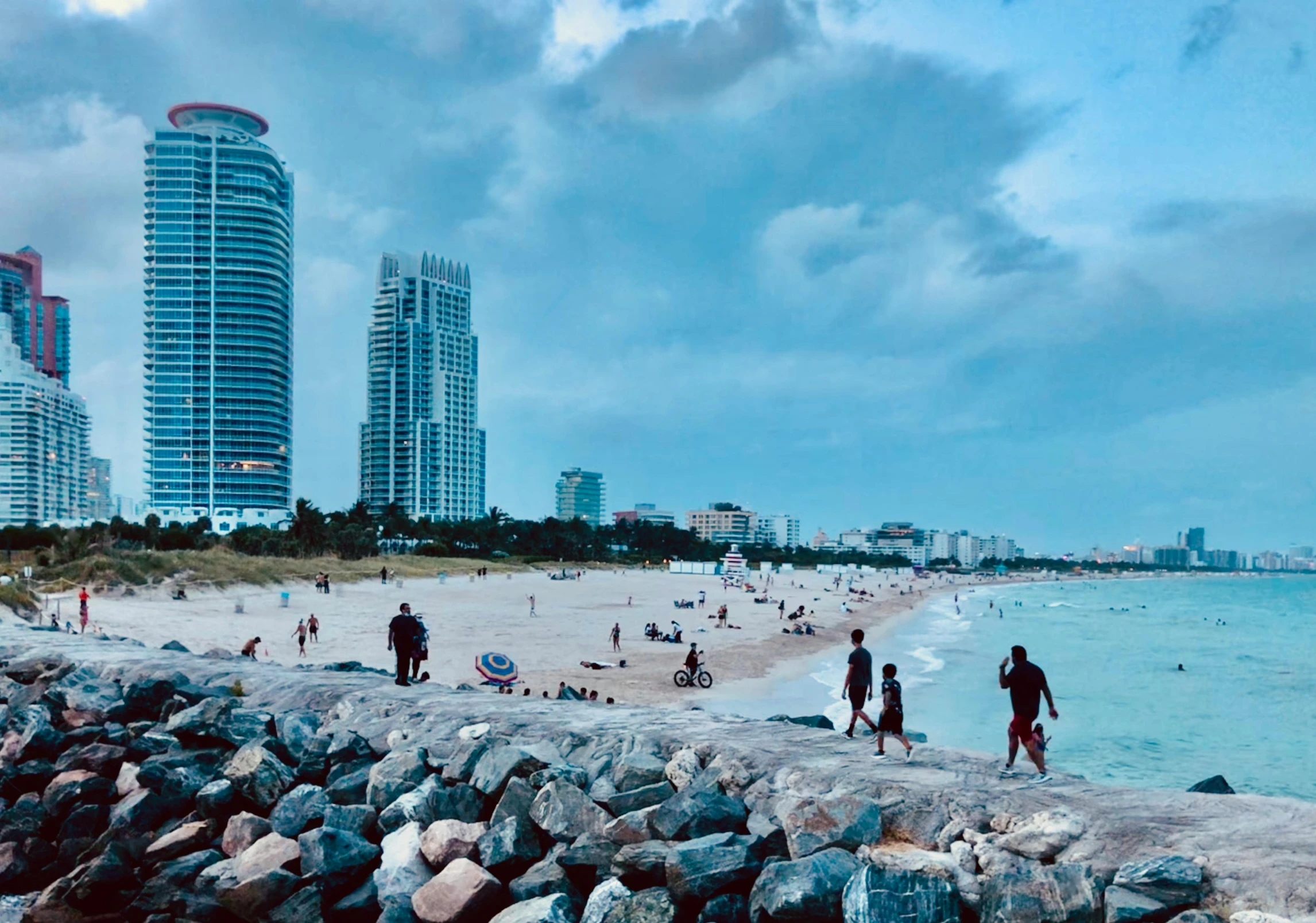 a group of people walking along a beach next to the ocean, by Niko Henrichon, unsplash contest winner, renaissance, palms and miami buildings, slide show, super wide view of a cityscape, youtube thumbnail