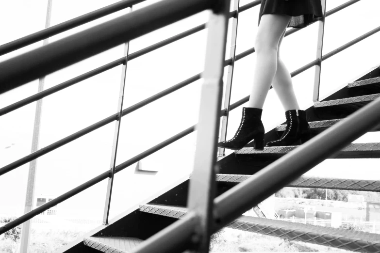 a black and white photo of a woman walking up a flight of stairs, a black and white photo, pexels, realism, black leather boots, wearing studded leather, 😭 🤮 💕 🎀, in motion