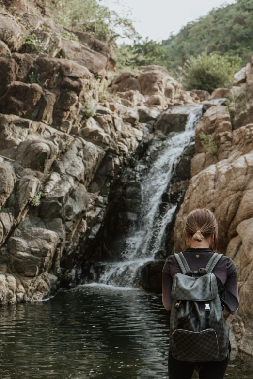 a woman standing in front of a waterfall, with a backpack, in a desert oasis lake, gaze down, streams and rocks