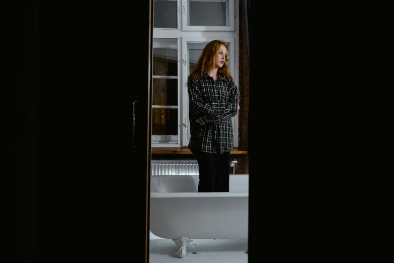a woman standing in a bathroom next to a bathtub, by Emma Andijewska, pexels contest winner, long shirt, redhead woman, dark clothing, mirroring