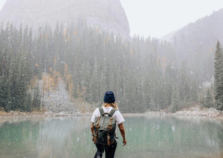 a person with a backpack standing in front of a lake, snow flurries, banff national park, foliage clothing, a person standing in front of a