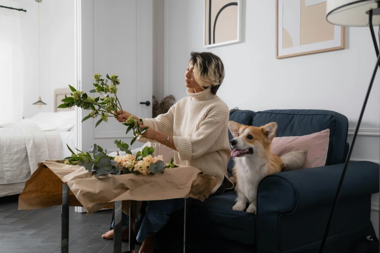 a woman sitting on a couch with a dog, a still life, by Emma Andijewska, pexels contest winner, picking up a flower, ikebana white flowers, cardboard, manuka