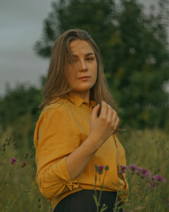 a woman standing in a field of tall grass, an album cover, by Attila Meszlenyi, pexels contest winner, color field, brown shirt, lovingly looking at camera, with yellow flowers around it, ((portrait))