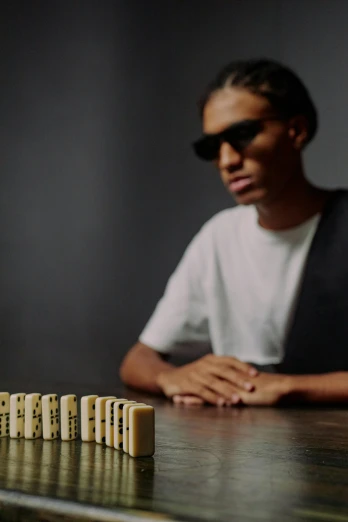 a man sitting at a table with dominos in front of him, an album cover, unsplash, visual art, ashteroth, gold glasses, in claymation, short depth of field