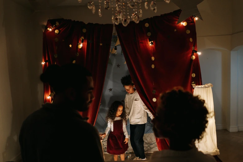 a couple of people that are standing in a room, inspired by The Family Circus, pexels contest winner, festive atmosphere, tiny girl looking on, red curtains, an escape room in a small