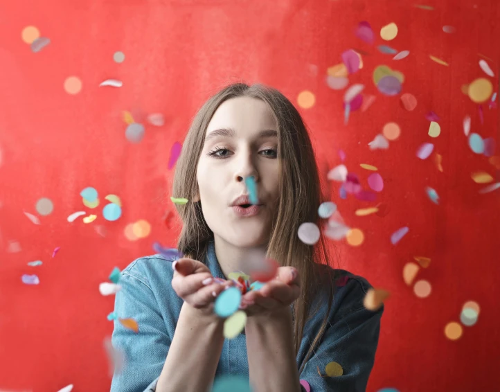 a woman blowing confetti in front of a red wall, promo image, multi colored, confetti, h3h3