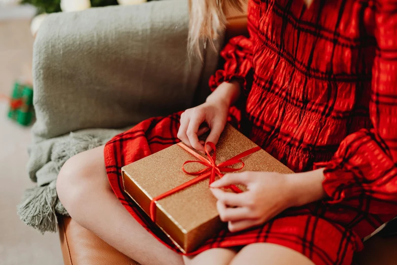 a woman sitting on a couch holding a present, by Julia Pishtar, pexels contest winner, wearing a red plaid dress, gold, closeup - view, ribbon