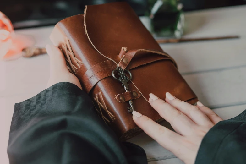 a person holding a brown book on top of a table, by Alice Mason, unsplash, wrapped in leather straps, luxury journal cover, lots of detail, crafts and souvenirs