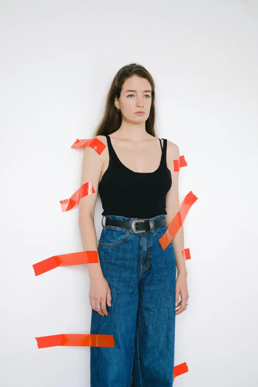 a woman standing in front of a wall covered in tape, by Matthias Stom, wearing a black cropped tank top, jeans and t shirt, wearing an orange t-shirt, labels