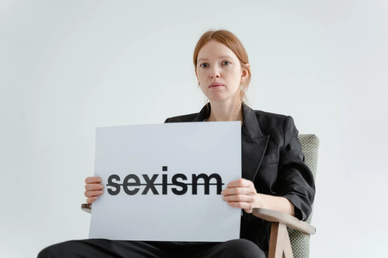 a woman sitting in a chair holding a sign that says sexism, a poster, trending on pexels, on a gray background, with pale skin, hr ginger, they are very serious