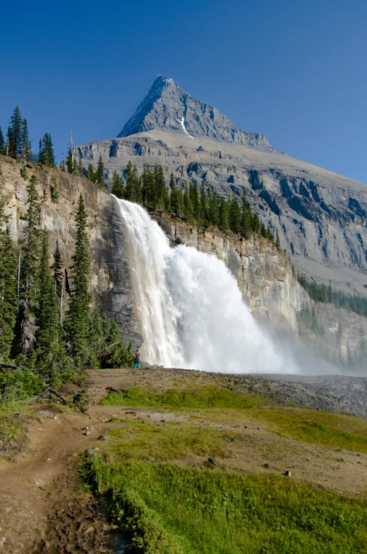 a man riding a bike down a dirt road next to a waterfall, banff national park, giant imposing mountain, slide show, information