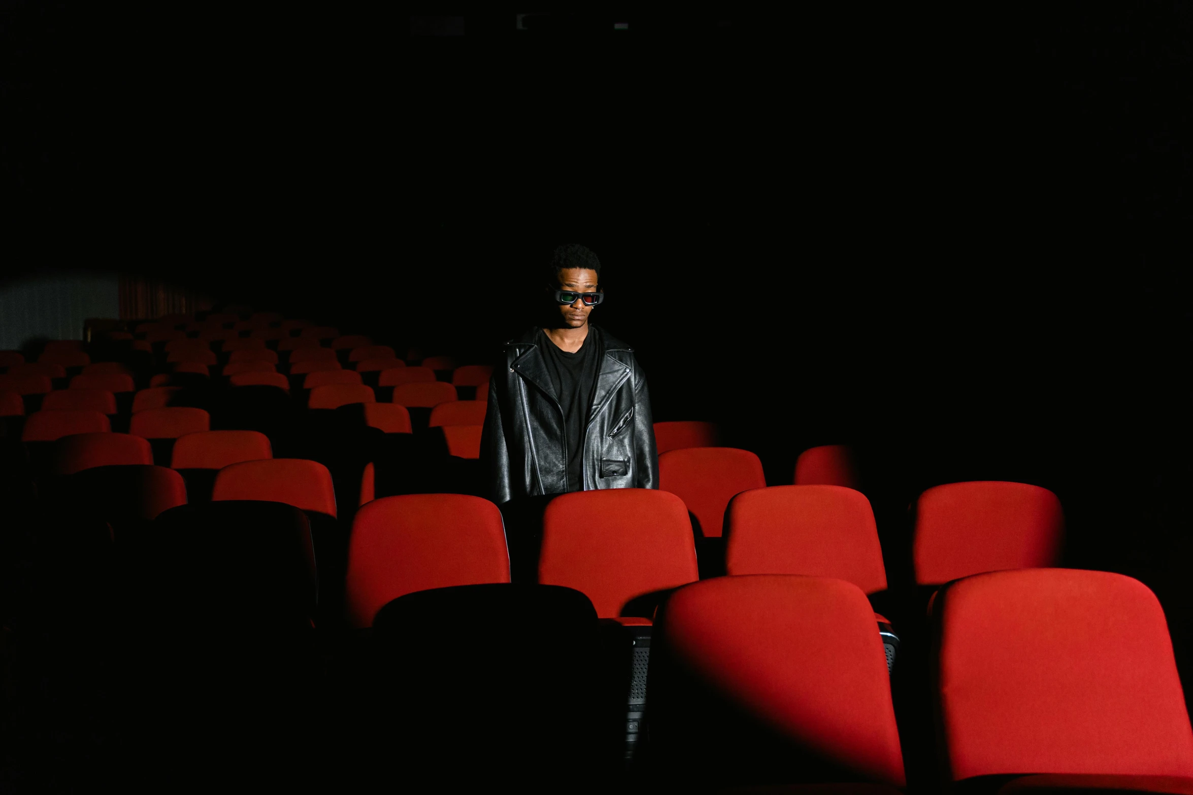 a man standing in front of rows of red chairs, an album cover, by James Morris, unsplash contest winner, sitting in a movie theater, ashteroth, dark shades, quality cinema model