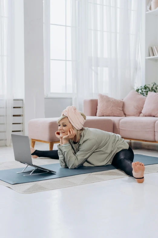 a woman laying on a yoga mat in front of a laptop, happening, curated collections, home video, wearing a pastel pink hoodie, placed in a large living room