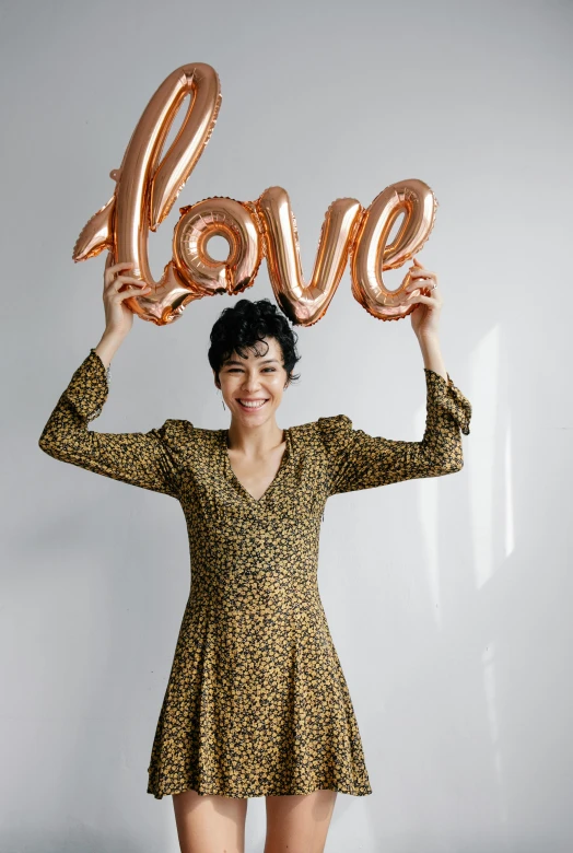 a woman holding up a love balloon in front of her face, curly pixie cut hair, wearing a dress of gossamer gold, official product photo, pose(arms up + happy)