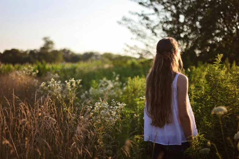 a woman standing in a field of tall grass, by Alice Mason, unsplash, girl with long hair, back lit, teenage girl, tiny girl looking on