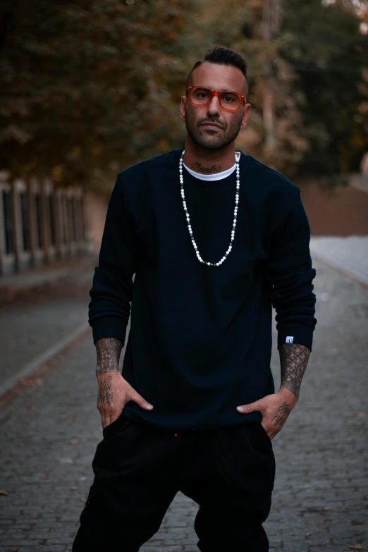 a man standing on a street with his hands in his pockets, inspired by Nadim Karam, unsplash, graffiti, wearing a dark sweater, wears a egyptian ankh necklace, in a navy blue sweater, album photo