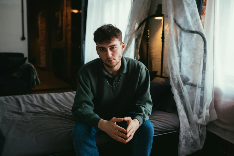 a man sitting on top of a bed next to a window, a portrait, pexels contest winner, 18 years old, hasbulla magomedov, wearing a green sweater, casey cooke