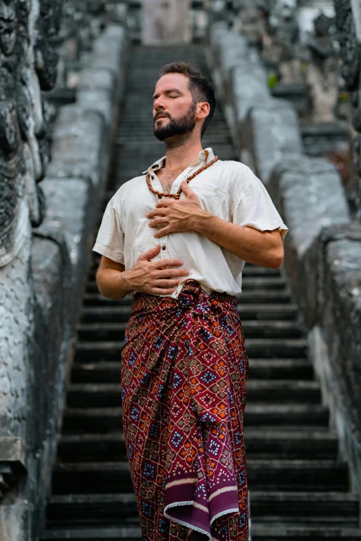 a man standing in front of a set of stairs, a portrait, inspired by I Ketut Soki, unsplash, renaissance, sarong, fantasy clothing, promotional image, brown