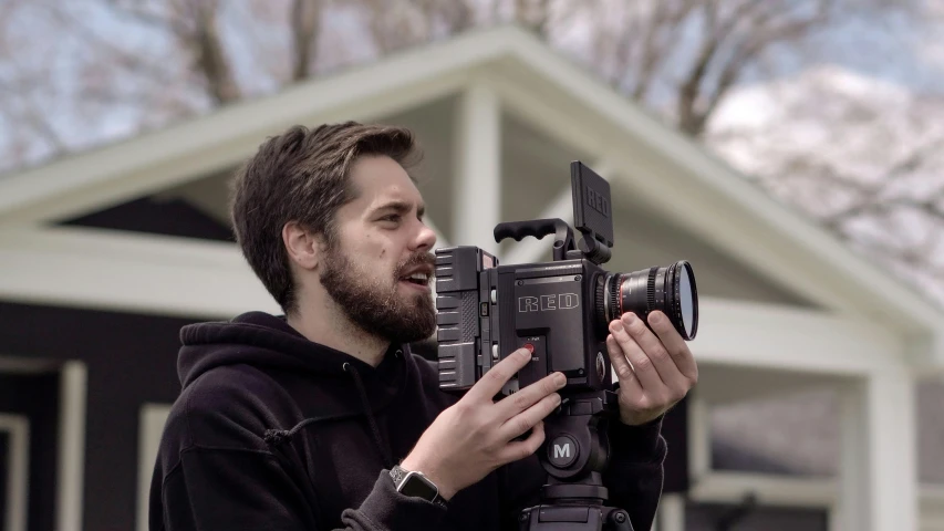 a man holding a camera in front of a house, red cinema camera, hasselblad quality, 8 k film still, mitchell mohrhauser