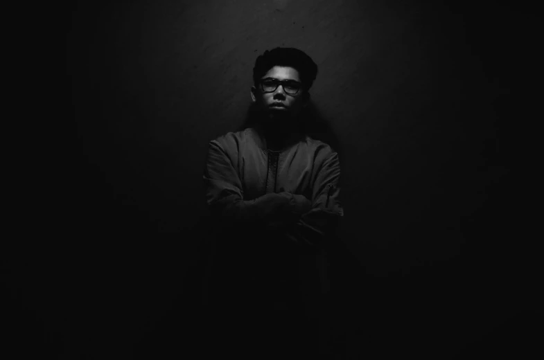 a man standing in the dark with his arms crossed, a black and white photo, by Youssef Howayek, unsplash, childish gambino, man with glasses, vantablack gi, brown skin man egyptian prince