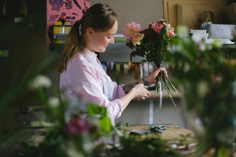 a woman standing in front of a bunch of flowers, a still life, process art, carefully crafted, in a workshop, friendly face, staff